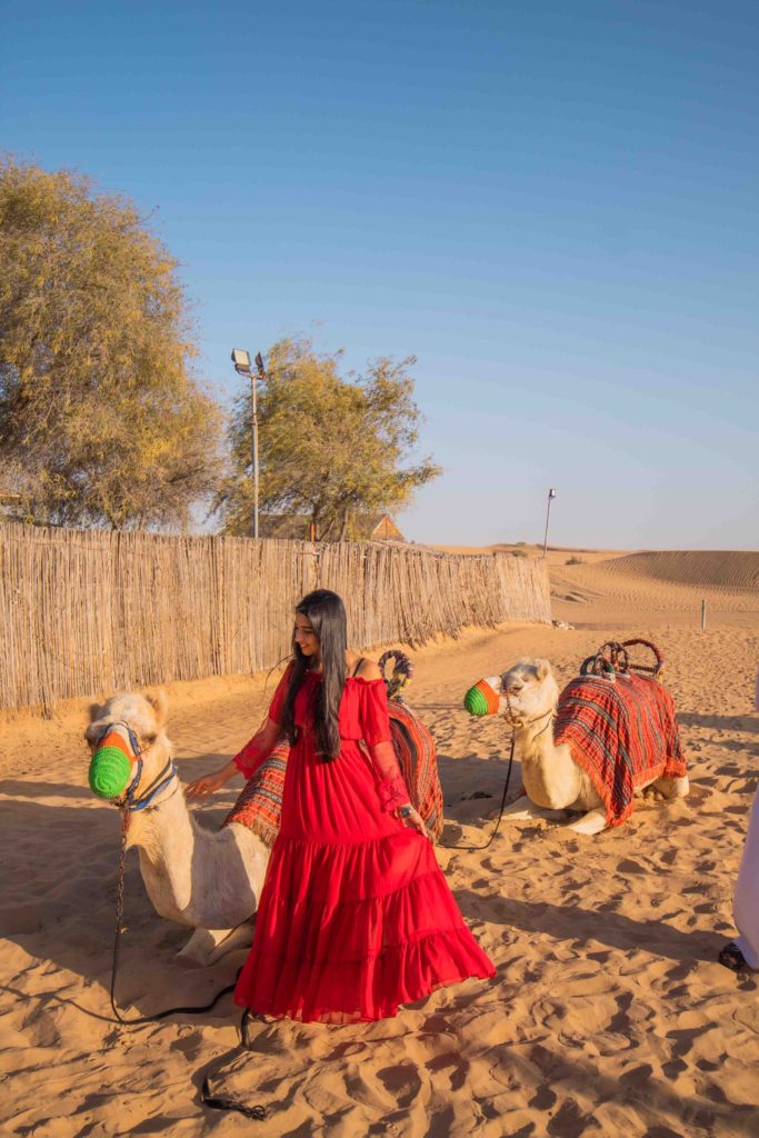 A girl in red dress standing with camels in desert