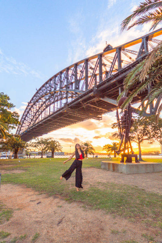 A girl in red top and black pants posing in front of Sydney harbour bridge at sunset