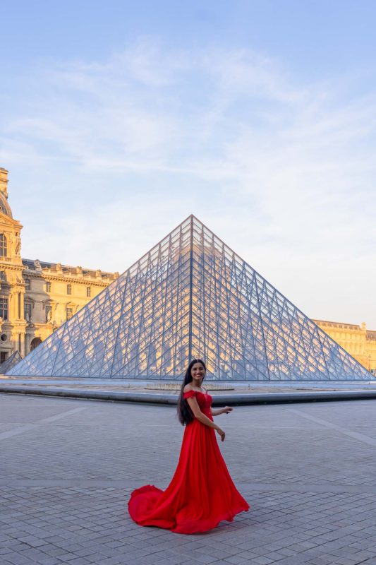 A girl in a red dress posing in front of the Louvre Pyramids.
