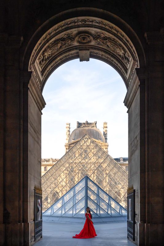 A girl in a red dress posing in front of the Louvre Pyramids.