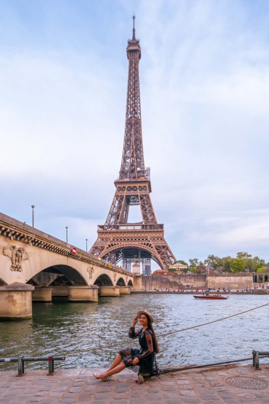 A girl in a black romper sitting at port Debilly with a view of the Eiffel tower